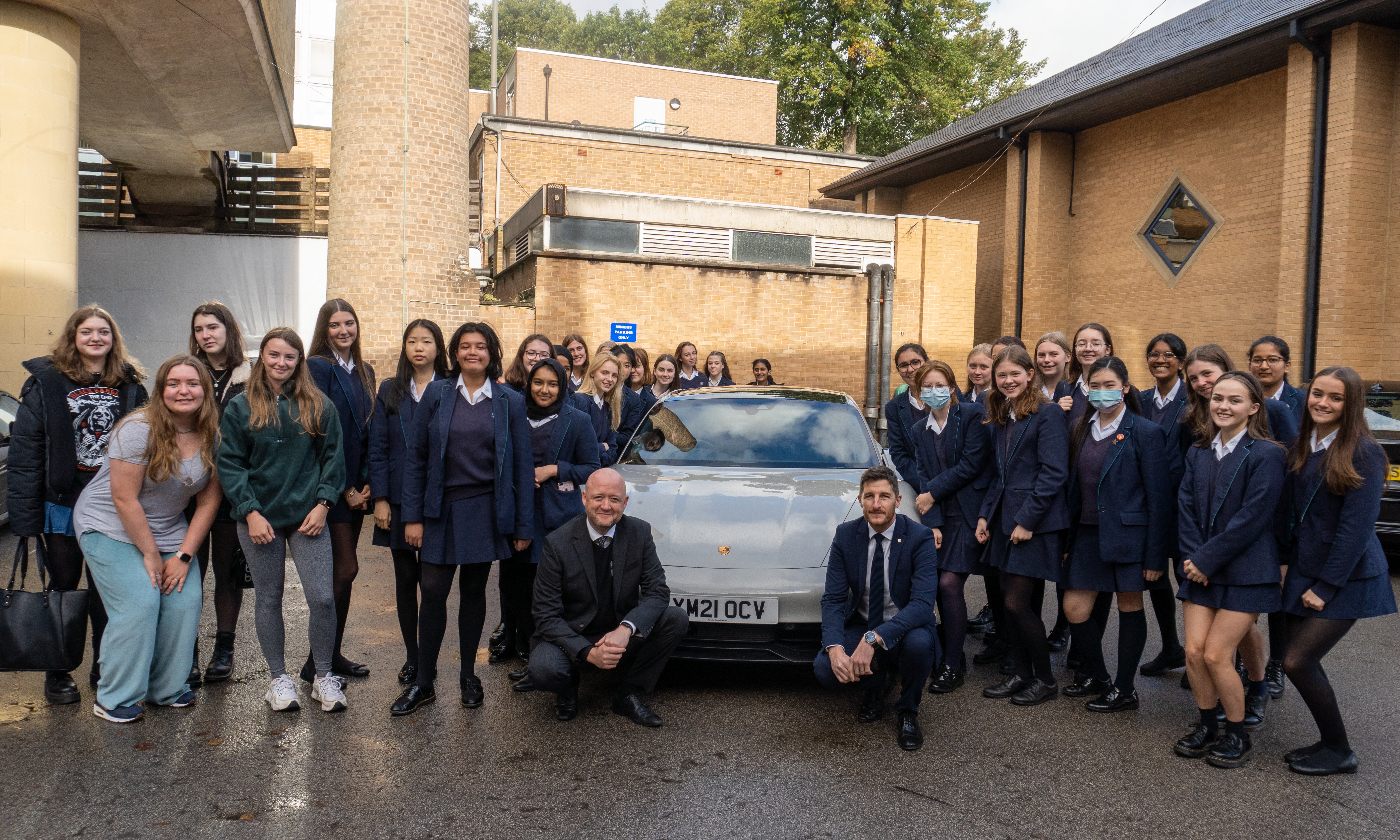 a group of girls in blue school uniform stood around a Porsche car with two men in suits at the front